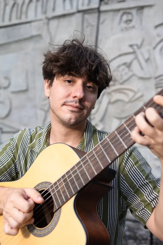 Leonel tocando violão em frente a um mural, na rua